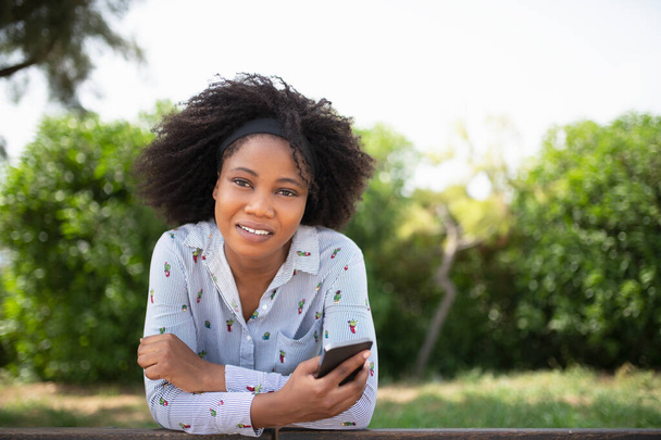Африканка дивиться на камеру і тримає мобільний телефон, використовуючи його в парку.. - Фото, зображення