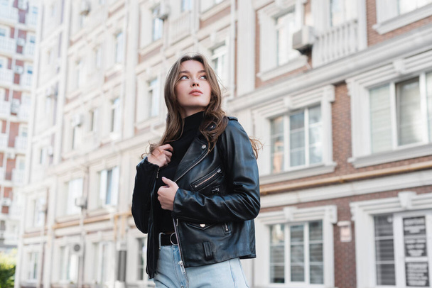 ヨーロッパの都会の通りにある黒いタートルネックとレザージャケットの若い女性  - 写真・画像