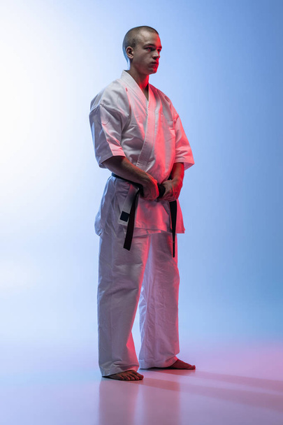 Zijaanzicht full-length portret van jonge professionele karate, judo sportman in wit kimono geïsoleerd over gradiënt wit blauwe achtergrond - Foto, afbeelding