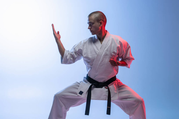 Retrato recortado de deportista caucásico de pie en una pose de lucha aislado sobre gradieta fondo azul blanco. Karate, judo, taekwondo sport - Foto, Imagen