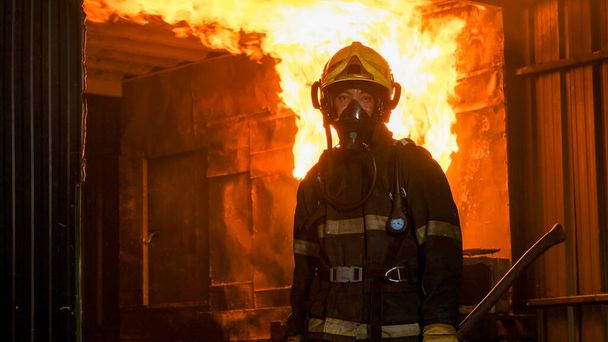Retrato de bombero con traje protector de pie delante del fuego en la pared de la cocina y mira a la cámara. También sostiene el hacha y se prepara para extinguir el fuego. - Foto, Imagen