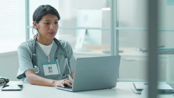 Gyönyörű spanyol doktornő gépel a laptopján, majd mosolyog a kamerába, miközben orvosi rendelőben dolgozik. - Felvétel, videó