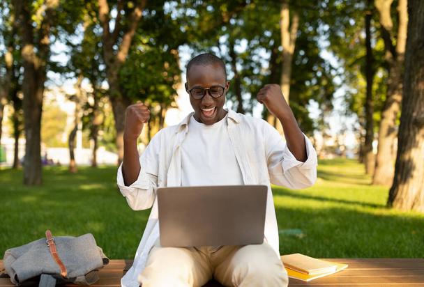 Η έννοια της εξ αποστάσεως εκπαίδευσης. Χαρούμενος μαύρος τύπος με φορητό υπολογιστή που κάνει χειρονομίες ΝΑΙ σε εξωτερικούς χώρους, κάθεται στην πανεπιστημιούπολη - Φωτογραφία, εικόνα