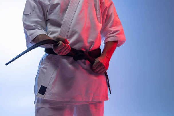 Cropped portrait of karate, judo sportsman in white kimono tying black belt isolated over gradient white blue background - Photo, Image
