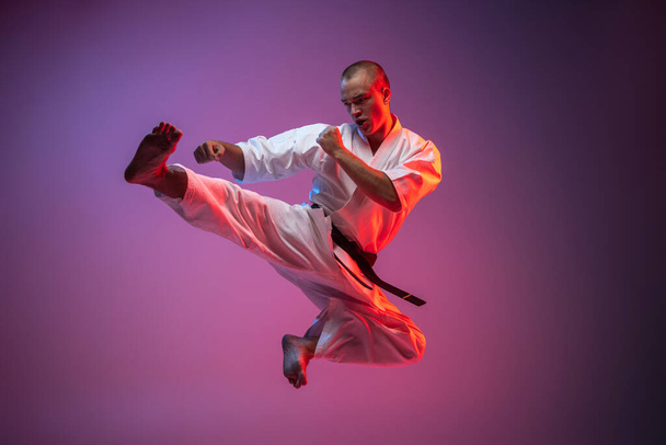 Młody wojownik ćwiczący latające kopnięcia na gradientowym różowym tle. Karate, judo, sport taekwondo - Zdjęcie, obraz