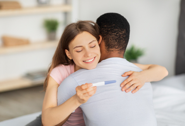 Joyful young woman hugging her black husband, holding positive pregnancy test at home - Foto, afbeelding