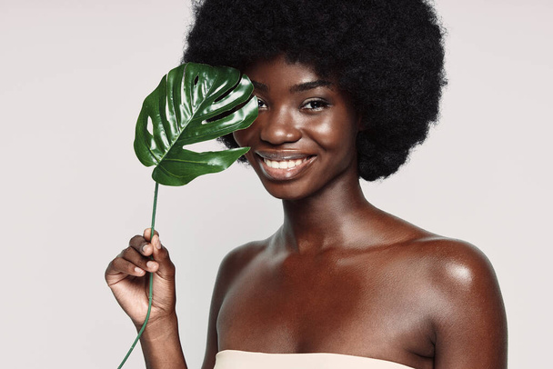Портрет красивой молодой африканской женщины, держащей растение возле лица и улыбающейся - Фото, изображение