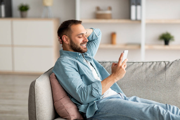 Przystojny tysiącletni facet relaksujący się na kanapie z telefonem komórkowym, uśmiechający się i piszący SMS-y do kolegi w domu - Zdjęcie, obraz