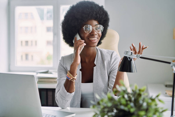 Beautiful young African woman talking on mobile phone and smiling while sitting at her working place in office - 写真・画像