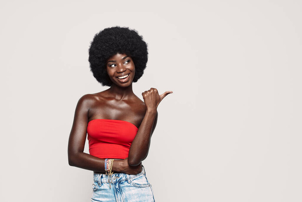 Beautiful young African woman pointing away and smiling while standing against gray background - Photo, Image