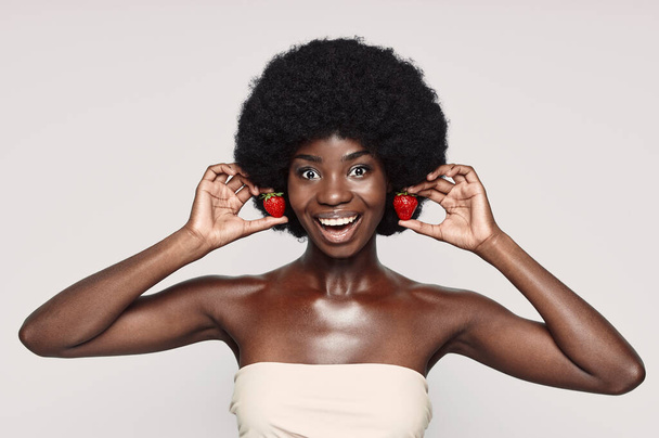 Portrait of beautiful young African woman holding strawberry like earrings and smiling - Foto, Imagem