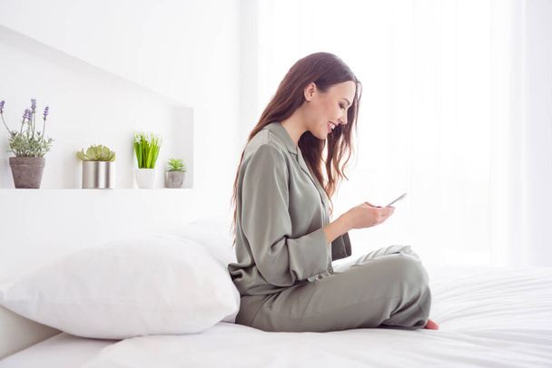 Profile side view portrait of attractive cheerful girl sitting on bed using device browsing at home house light white interior - Фото, зображення