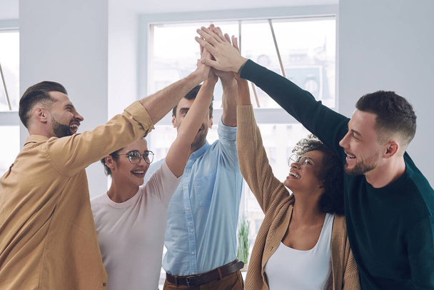 Eine Gruppe fröhlicher junger Leute in schicker Freizeitkleidung hält die Hände zusammen und lächelt beim Treffen im Büro - Foto, Bild