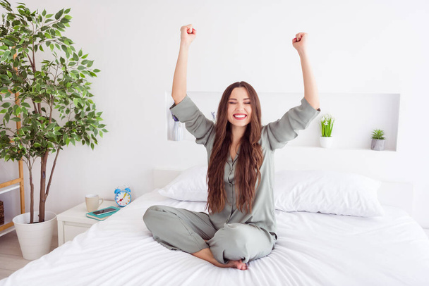 Photo of adorable lucky young lady grey sleepwear sitting bed smiling rising fists indoors room home - Foto, Imagem
