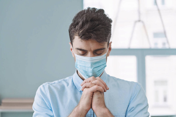 Handsome young man in protective face mask praying with his hands clasped and eyes closed - Foto, Imagem