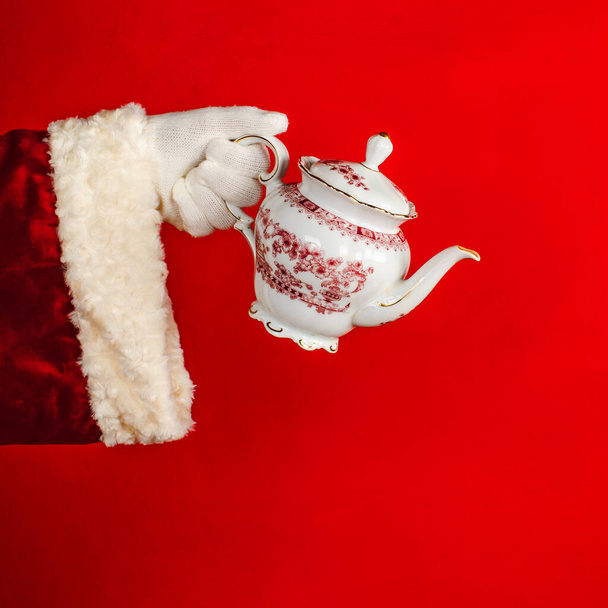 Die Hand des Weihnachtsmannes hält eine Porzellanteekanne mit Mustern. Weihnachtsurlaub, Wintertradition Heißgetränk. - Foto, Bild