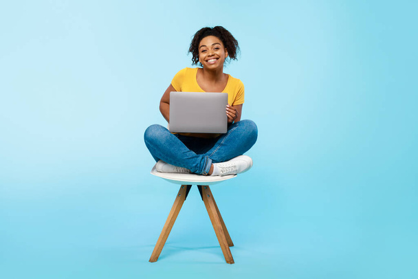 Joven mujer negra alegre que trabaja en línea, sentado en la silla y usando el ordenador portátil en fondo azul del estudio, longitud completa - Foto, imagen