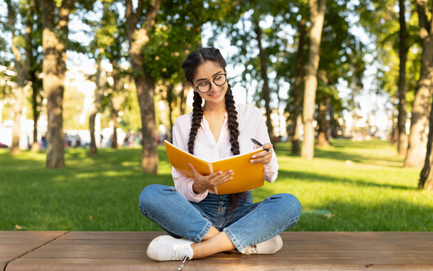 Nainen intialainen opiskelija tyttö kirjoittaa muistiinpanoja kannettavan istuessaan penkillä yliopiston kampuksella tai puistossa - Valokuva, kuva