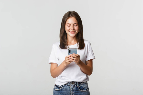 Χαμογελώντας ελκυστική κοπέλα χρησιμοποιώντας το κινητό τηλέφωνο και κοιτάζοντας την οθόνη ευχαριστημένος - Φωτογραφία, εικόνα