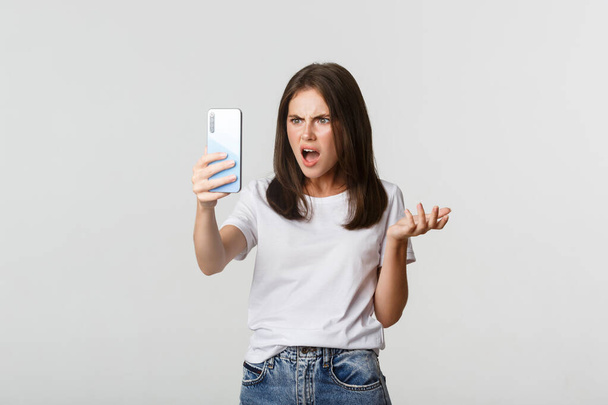 Turhautunut nuori nainen, jolla on argumentti videopuhelu, tilalla älypuhelin, pysyvä valkoinen tausta - Valokuva, kuva