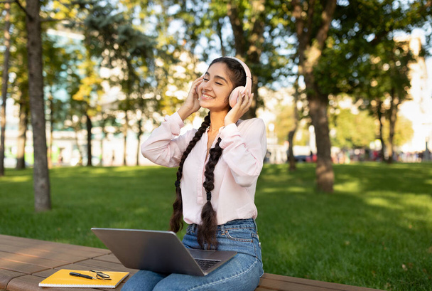 Iloinen intialainen opiskelija tyttö lepää kampuksella kannettava tietokone, yllään kuulokkeet ja kuuntelee musiikkia, istuu penkillä - Valokuva, kuva