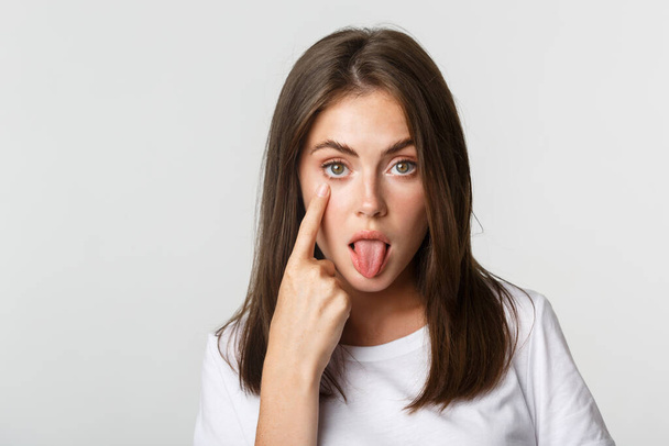 Close-up of silly attractive young girl showing tongue and pulling eyelid, watching you - Foto, afbeelding