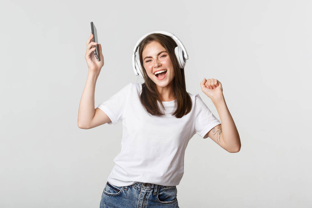 Happy beautiful brunette girl dancing and listening music in wireless headphones, holding smartphone - Photo, Image