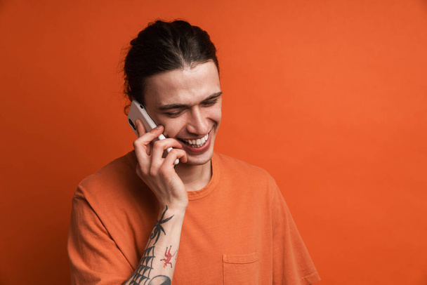 Young white man with tattoo laughing while talking on cellphone isolated over orange background - Foto, imagen