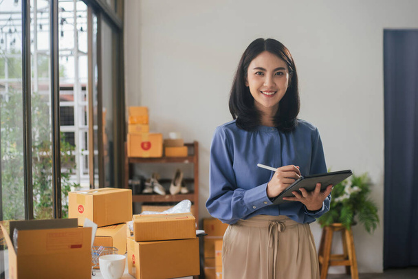 Portrait business Asian woman smiling look at camera and use tablet checking information on parcel shipping box before send to customer. Entrepreneur small business working at home. - Photo, Image