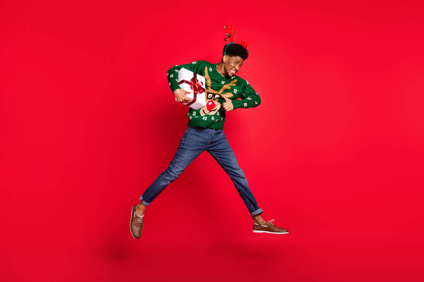 Photo of guy jump run carry x-mas present raise thumb up wear green deer sweater headband isolated on red background - Foto, Imagem