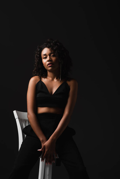sexy african american woman in satin crop top sitting on white chair isolated on black  - Photo, Image