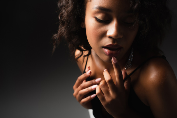 соблазнительная молодая африканская американка в сексуальном топе, касаясь губ, позируя на черном  - Фото, изображение