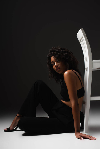 giovane donna afroamericana in sexy crop top e pantaloni seduti vicino sedia bianca mentre posa sul nero  - Foto, immagini