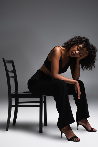 full length of brunette african american woman in stylish outfit sitting on chair on grey - Foto, immagini