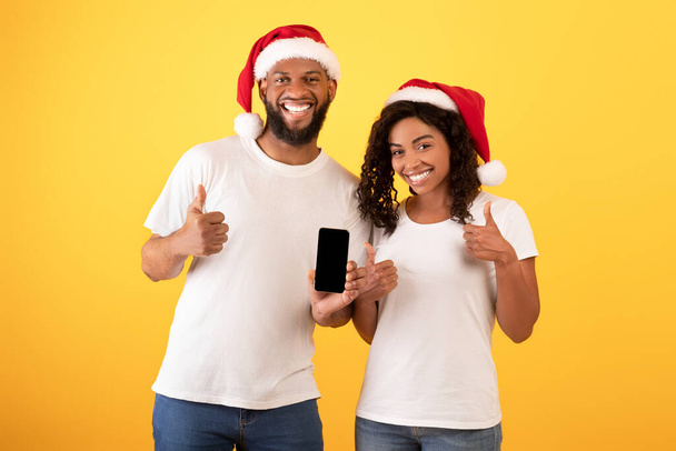 Nowa strona. Szczęśliwi czarni małżonkowie trzymający smartfona z pustym ekranem i pokazujący kciuki w górę, żółte tło - Zdjęcie, obraz