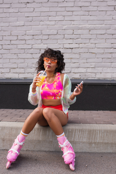 volledige lengte van stijlvolle Afrikaanse Amerikaanse vrouw in zonnebril en rolschaatsen drinken van sinaasappelsap en met smartphone  - Foto, afbeelding
