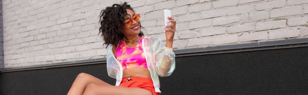 smiling african american woman in sunglasses using smartphone outside, banner - Foto, afbeelding
