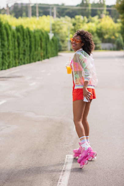pleine longueur de femme afro-américaine heureuse patinant sur patins à roulettes tout en tenant une tasse en plastique avec du jus d'orange et un téléphone portable - Photo, image