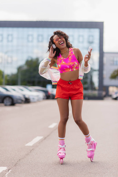 cheerful african american woman in wireless headphones and on roller skates singing while holding smartphone  - Fotoğraf, Görsel