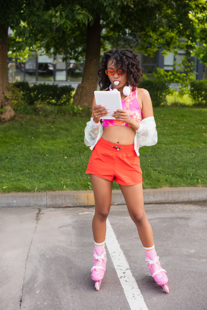 mujer afroamericana rizada en auriculares inalámbricos y en patines de ruedas que soplan goma de mascar mientras usa la tableta digital afuera  - Foto, imagen