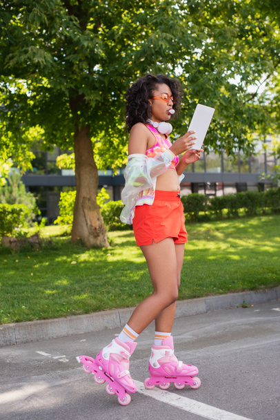 curly african american woman in headphones and on roller skates blowing bubble gum while using digital tablet outside  - Photo, Image