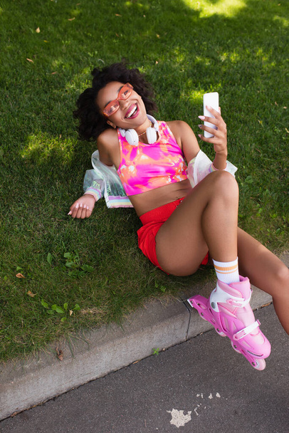 cheerful african american woman on roller skates smiling while holding smartphone and resting on grass - Φωτογραφία, εικόνα