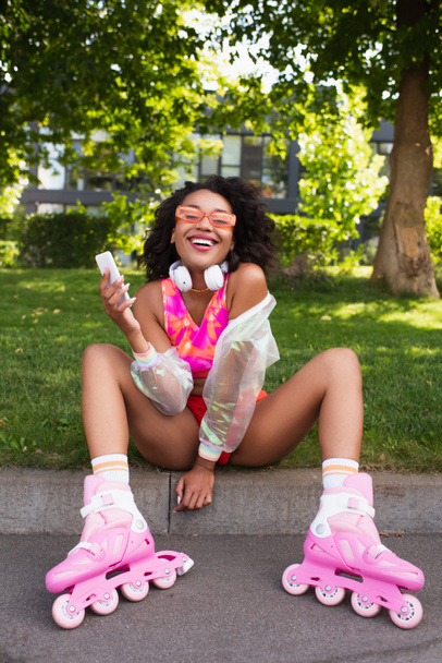 happy african american woman in headphones and on roller skates smiling while holding smartphone  - 写真・画像