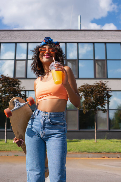 Mutlu Afro-Amerikalı kadın başörtüsü takmış elinde portakal suyu ve dışarıda uzun pano olan plastik bir bardak tutuyor.  - Fotoğraf, Görsel