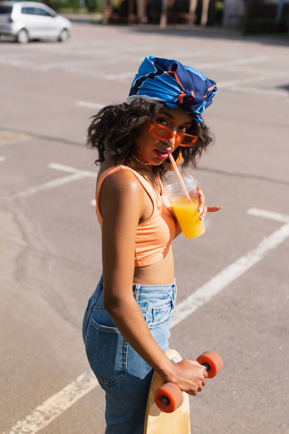 νεαρή Αφροαμερικανή γυναίκα με γυαλιά ηλίου που πίνει χυμό πορτοκαλιού και κρατά το longboard έξω  - Φωτογραφία, εικόνα