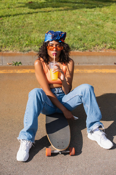 mulher africana americana em óculos de sol segurando copo de plástico e beber suco de laranja enquanto sentado no longboard fora  - Foto, Imagem