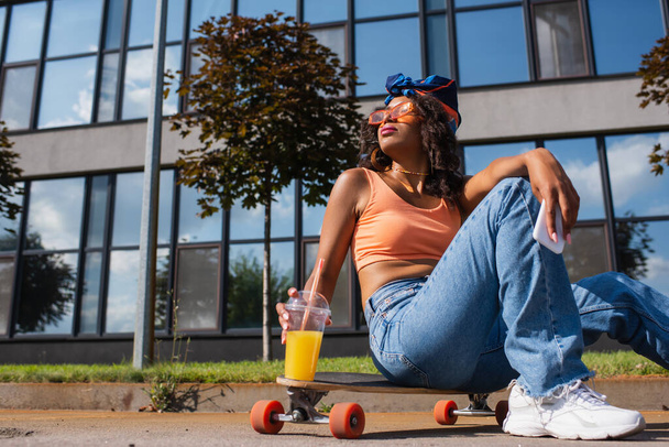 african american woman in sunglasses sitting on longboard while holding smartphone and plastic cup  - Photo, Image