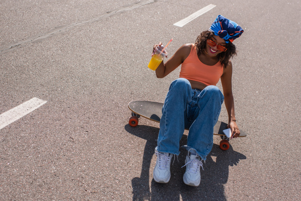 mulher americana africana feliz em óculos de sol sentado no longboard enquanto segurando smartphone e copo de plástico  - Foto, Imagem