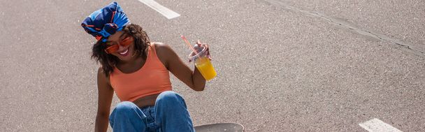 fröhliche afrikanisch-amerikanische Frau mit Sonnenbrille, Plastikbecher mit Orangensaft in der Hand und draußen auf Longboard sitzend, Banner - Foto, Bild
