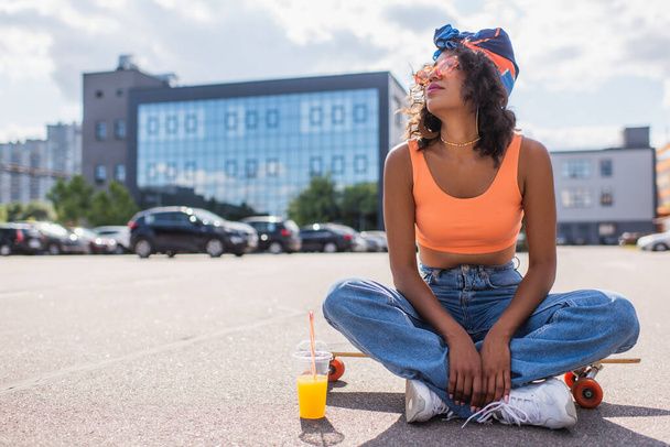 mulher africana americana em óculos de sol sentado com pernas cruzadas em longboard perto de copo de plástico com suco de laranja no asfalto  - Foto, Imagem